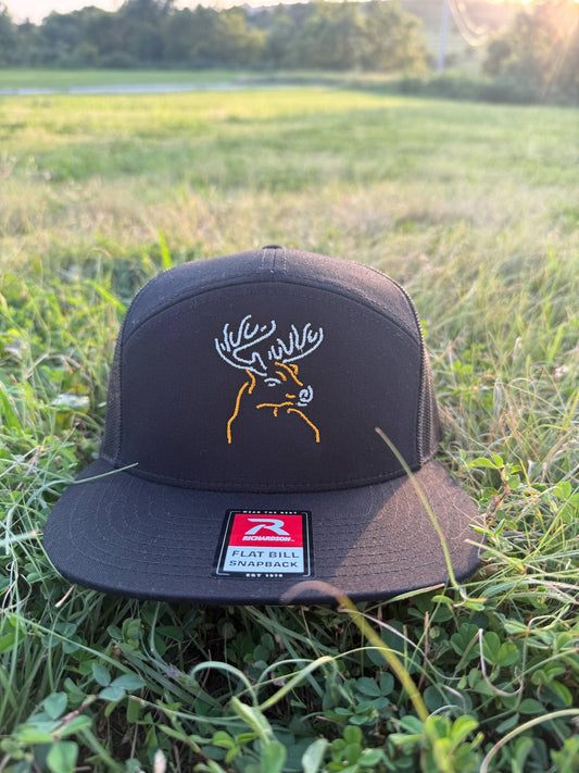 Black snapback hat featuring neon-style glowing buck embroidery on front crown, named Neon Buck, placed on grassy field at sunset.
