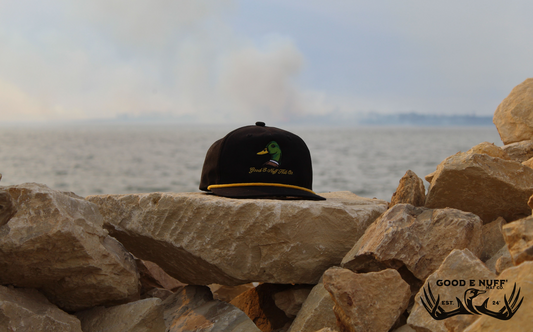 Black hat with embroidered mallard design and "Good E Nuff' Hat Co." text, displayed on rocks near water.