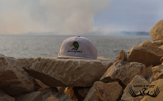 Grey "King of the Pond" hat with mallard embroidery on rocks by water.