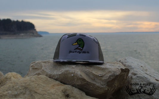 Mallard embroidered Richardson 7-panel hat with camo bill and mesh back on rocks by a lake.