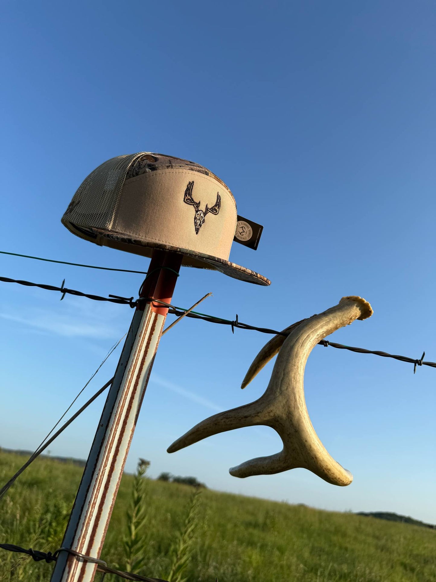 Dead Head tan and camo hat with whitetail skull design hanging on fence post outdoors