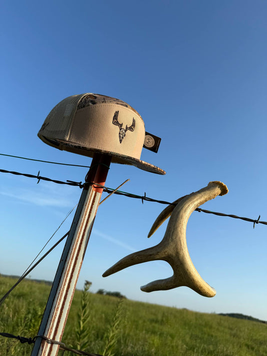 Dead Head tan and camo hat with whitetail skull design hanging on fence post outdoors