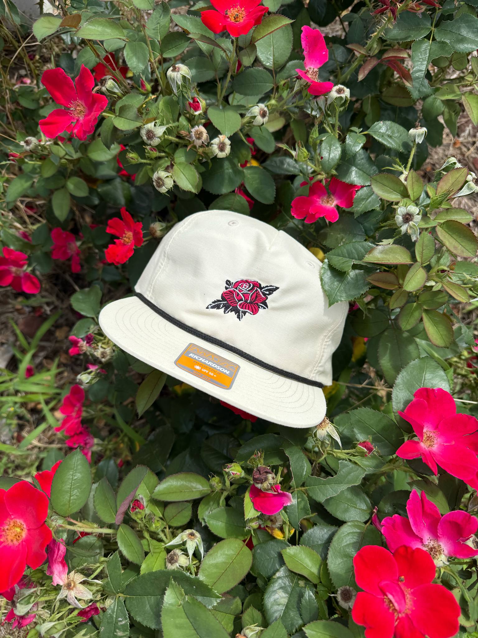 Cream-colored Richardson cap with embroidered rose and black rope detail among red flowers.