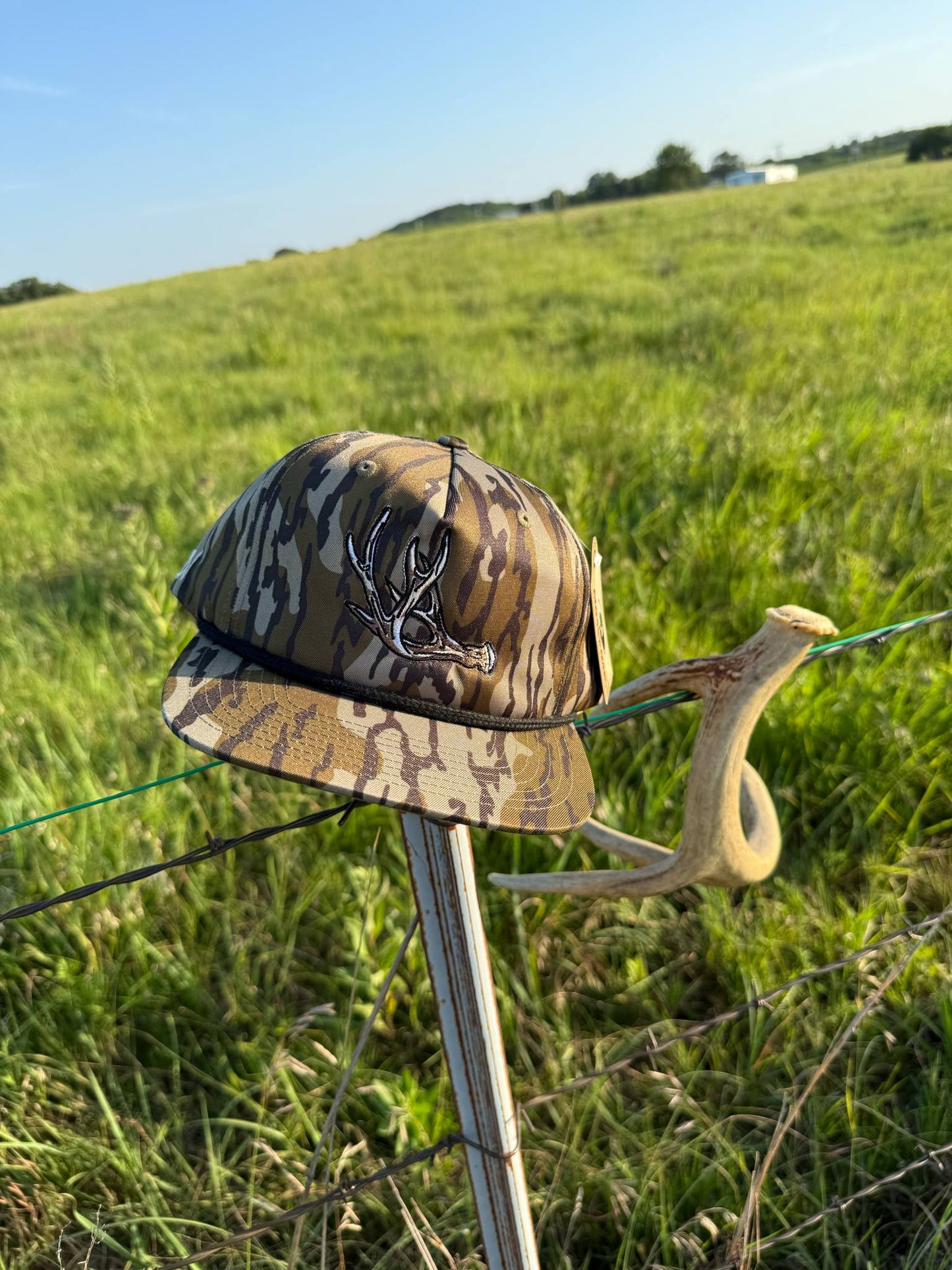 Shed Hunter camo hat with antler embroidery resting on a fence post in a grassy field.