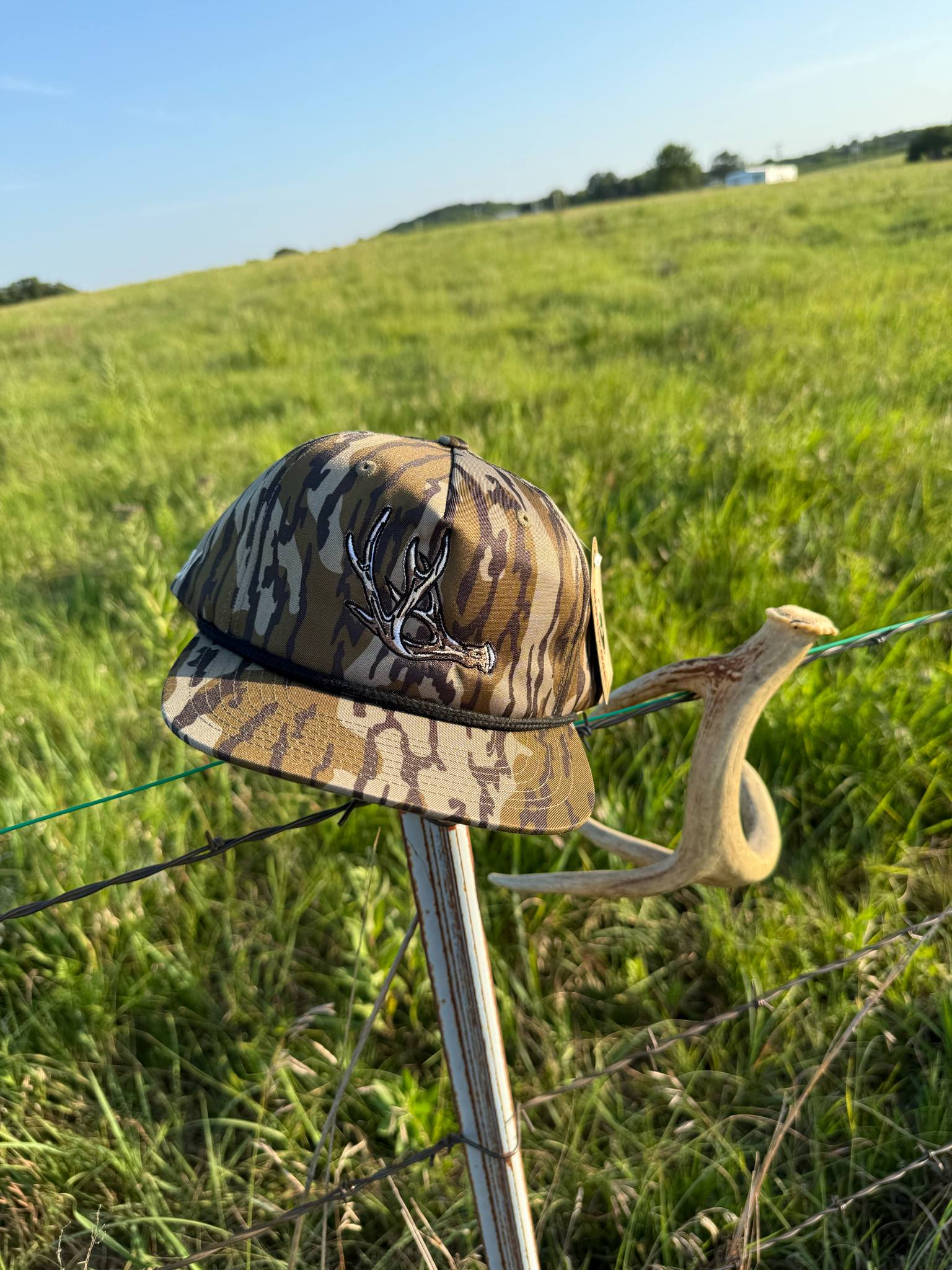 Shed Hunter camo hat with antler embroidery resting on a fence post in a grassy field.