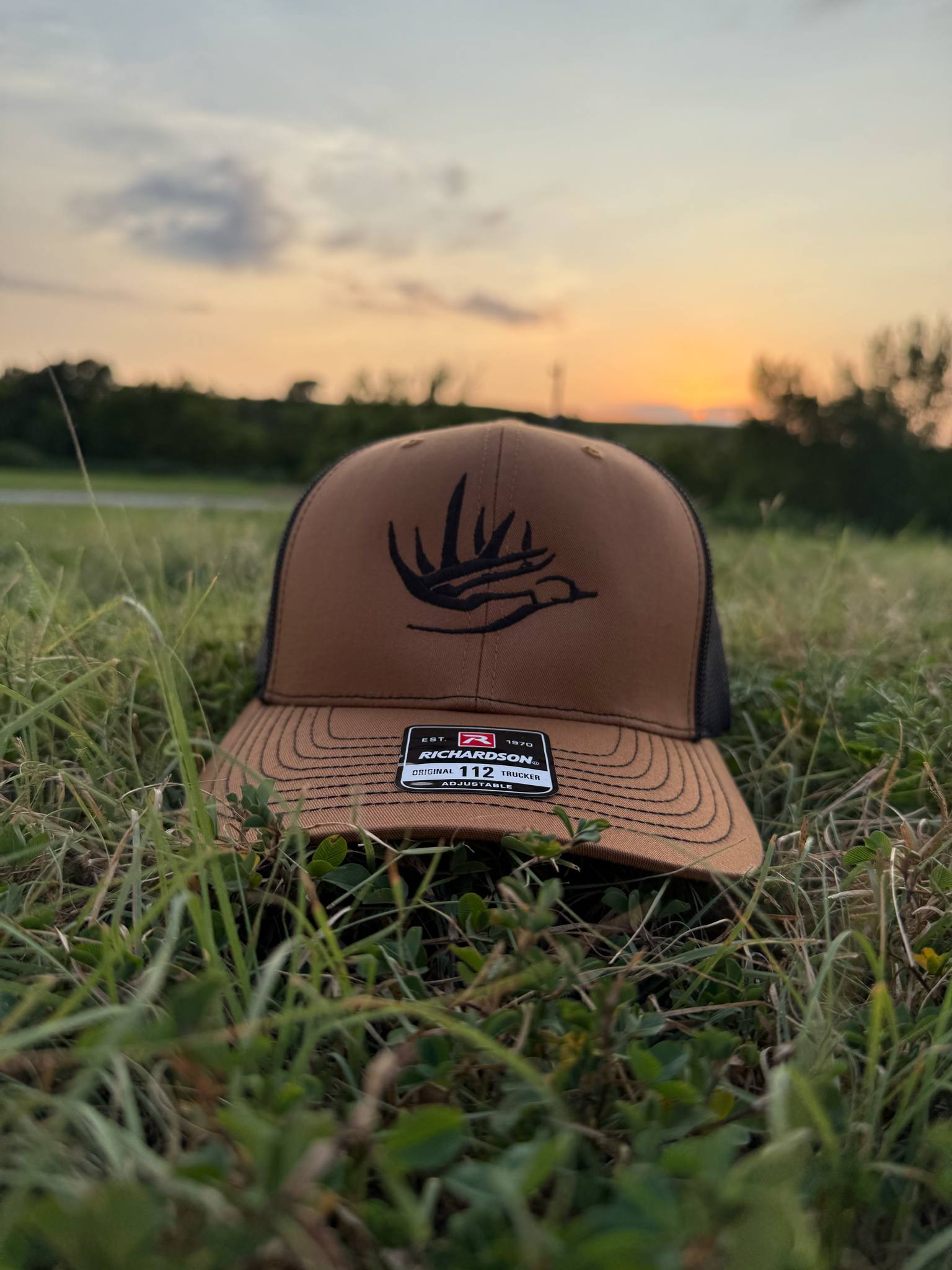The Roamer brown and black Richardson 112 hat with antler and duck graphic on grass at sunset