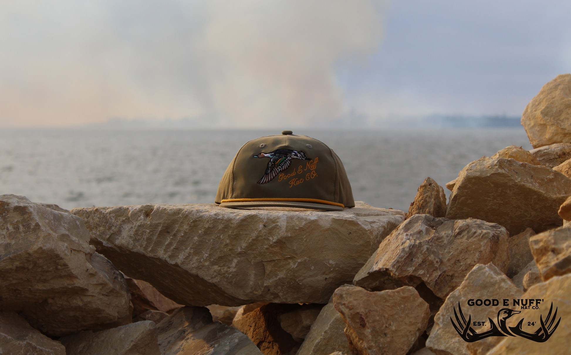 Old School Pintail hat with embroidered duck and "Good E Nuff' Hat Co." text on an olive base with retro rope detail, set on rocks by a lake.