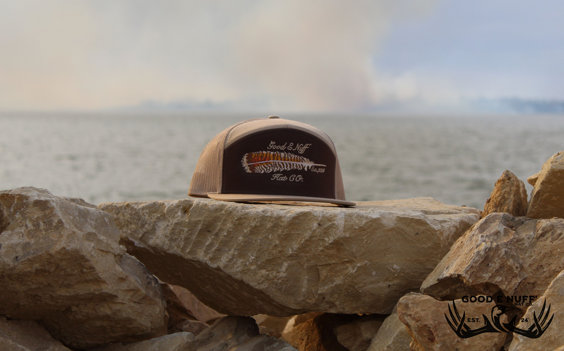 Rustic "The Roost" hat with turkey feather embroidery on rocks by the water.
