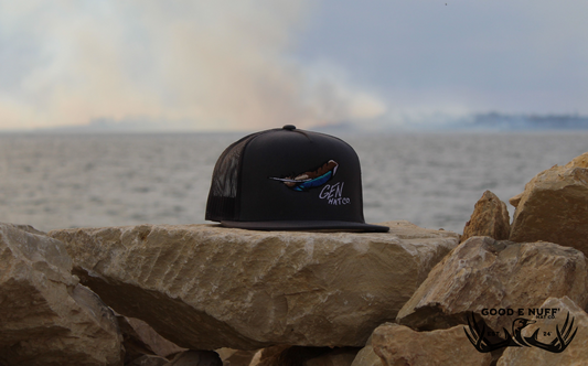 Grey trucker hat with blue-wing teal feather embroidery, GEN Hat Co. script, flat bill, mesh back, on rocky shore.