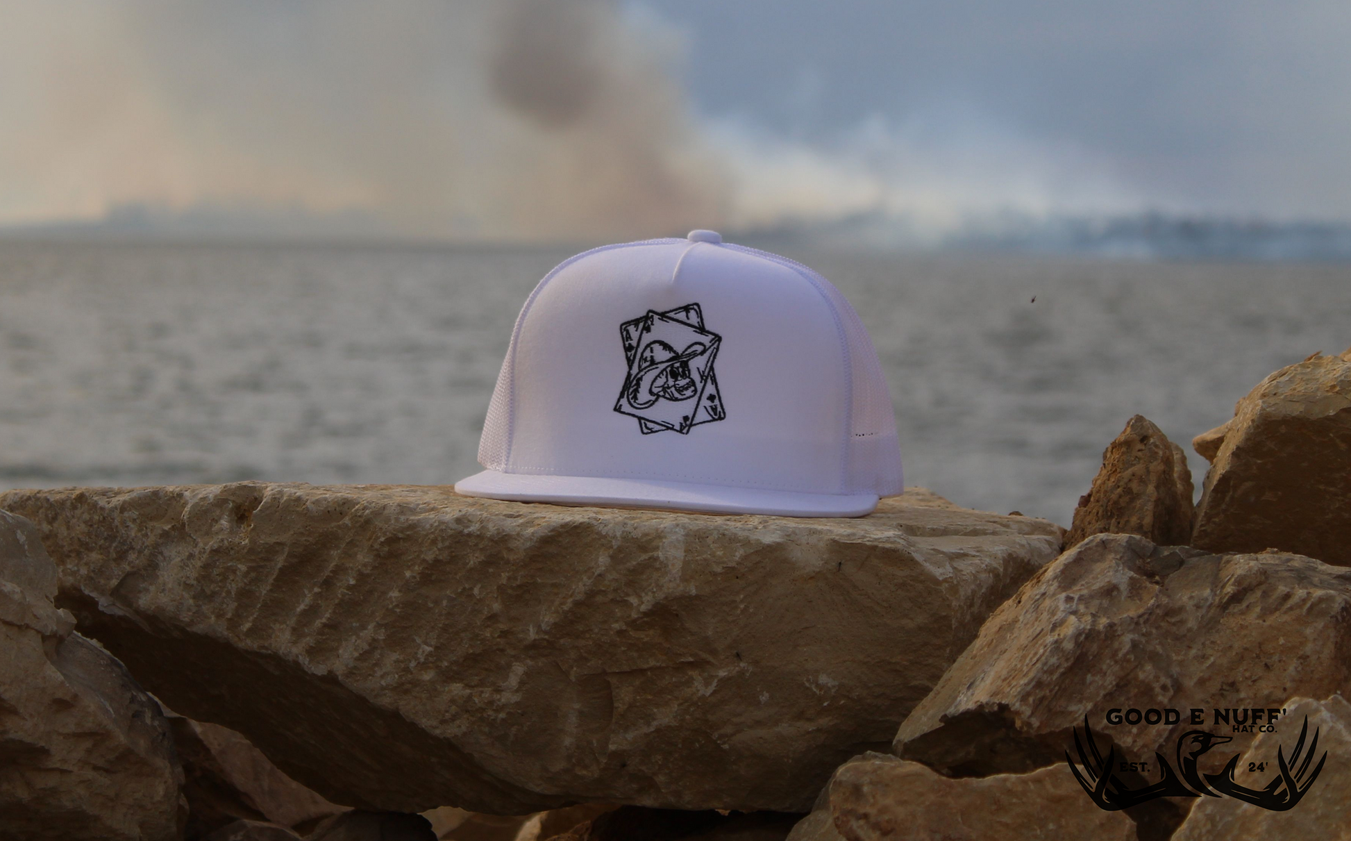 Cowboy skull embroidered "Pocket Aces" hat with white front and mesh back on rocks by the water.